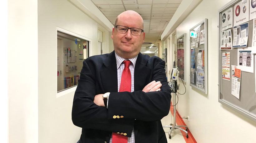 Professor John Heath stands in a hospital hallway, smiling, with his arms folded confidently.