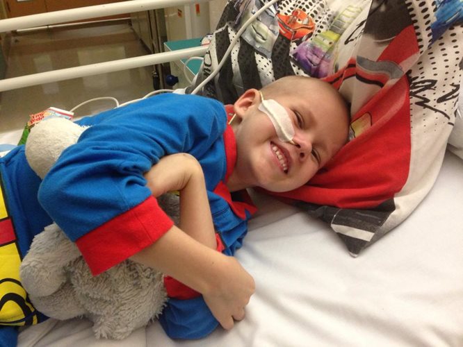 Zach is smiling while lying on a hospital bed, wearing a superhero costume and hugging a teddy-bear.