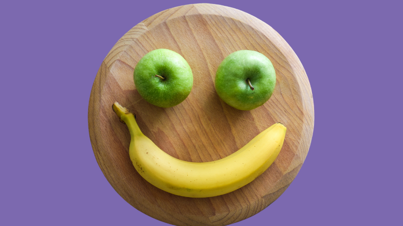 On a purple backdrop sits a round chopping board with a banana and two green apples arranged like a smiley face.
