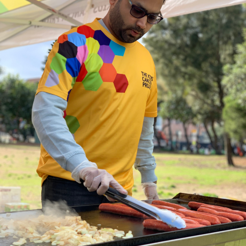 A man wearing a gold TKCP tee-shirt is cooking sausages and onions on a BBQ hotplate.