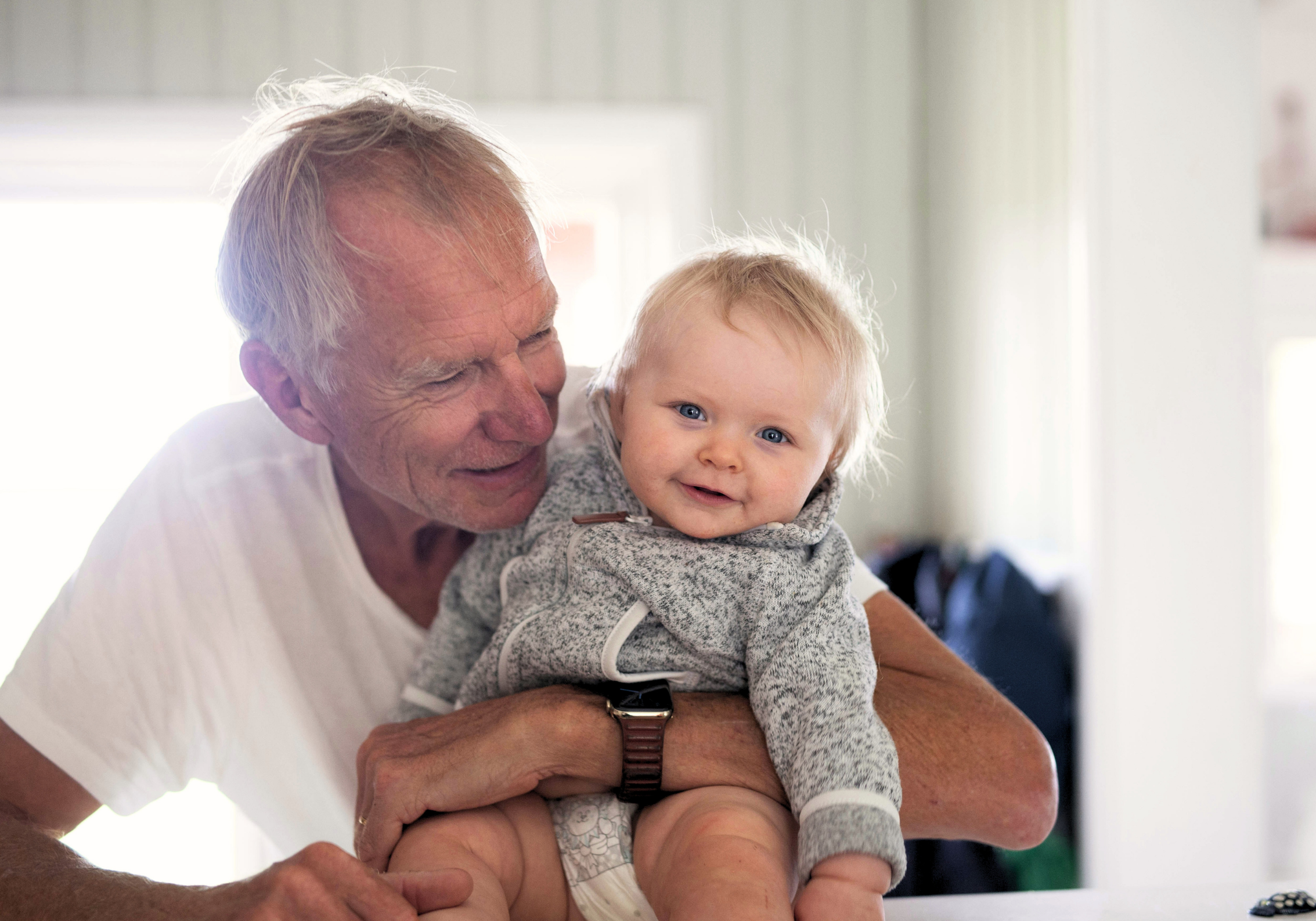 Grandfather holding baby grandson, looking at him and smiling while the baby smiles to the camera