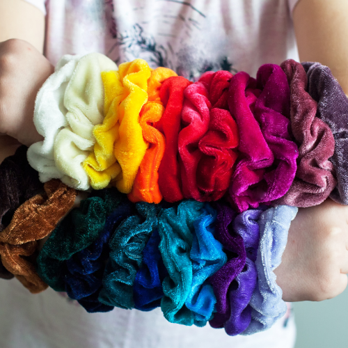 A person's forearms are fully covered in bright, colourful, velvet scrunchies, worn like bracelets.