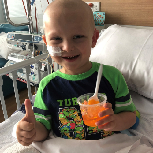 Ollie sitting up in his hospital bed with a half eaten cup of orange jelly giving a thumbs up