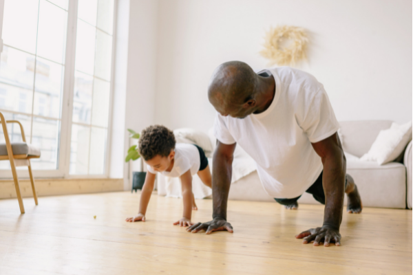 A father and his young son do some push-ups together in their living room.