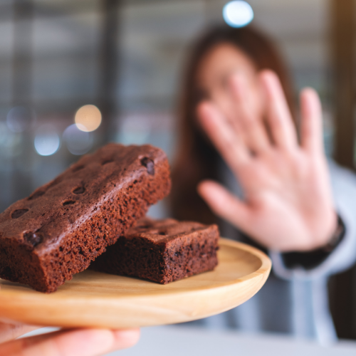 In the foreground is a plate of delicious brownies, the background is blurred, but depicts a person holding up their hand to refuse the brownies.