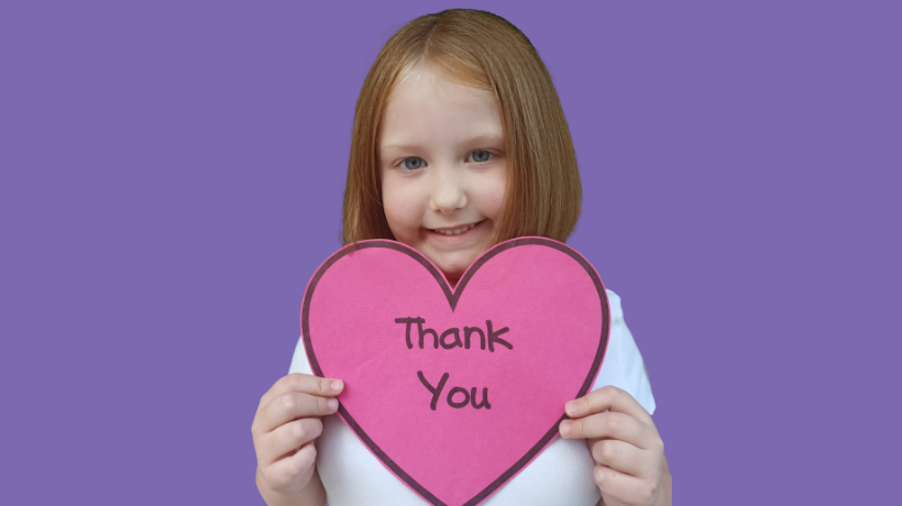 Imogen is smiling in front of a purple backdrop and holding a pink, paper heart with the words 