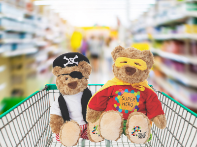 A bear dressed like a pirate and a bear dressed like a superhero are sitting in a shopping trolley; the background is a blurred supermarket aisle.