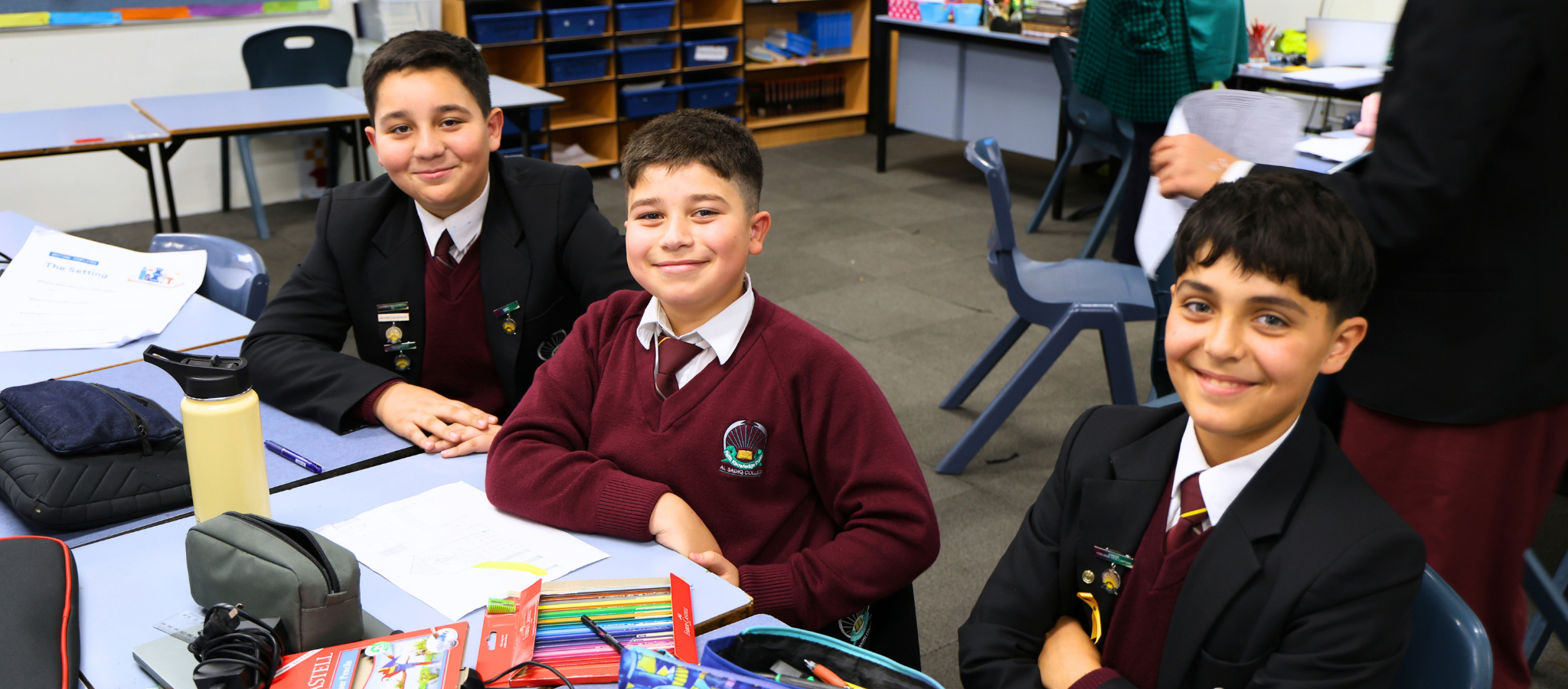 Three school boys sat at a table in a classroom, smiling at the camera.