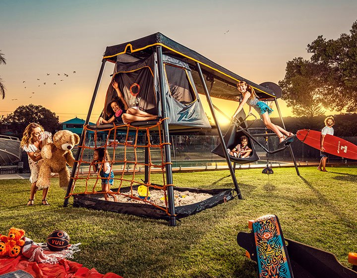 Vuly outdoors play equipment set up in a grassy area at sunset, children are playing happily under supervision; there is a picnic set up at the bottom left corner of the picture.