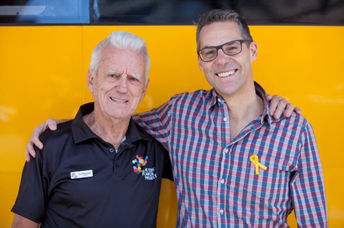 Professor Ricky Johnstone smiles arm-in-arm with Col Reynolds in front of a yellow backdrop.