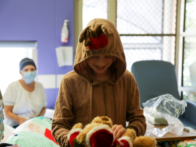 A small child wearing a Rudolf onesie smiles while examining a teddy bear dressed like Santa Claus.