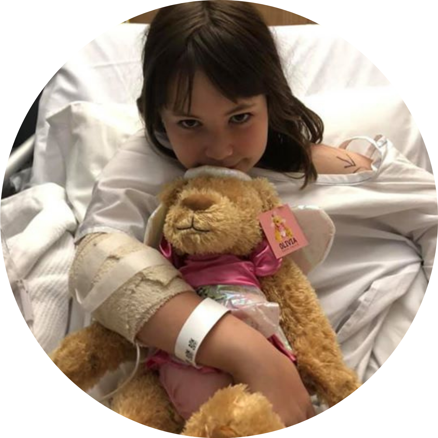 A little girl in a hospital bed is hugging a donated bear that is dressed in a pink angel costume.