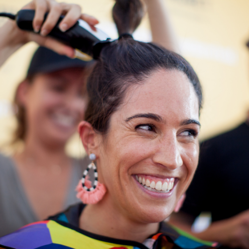 A smiling woman wearing a TKCP cape is getting her hair shaved off to raise money for kids' cancer research.
