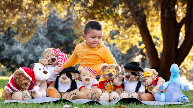 CJ is happily sitting on a picnic rug in a park, surrounded by TKCP bears.