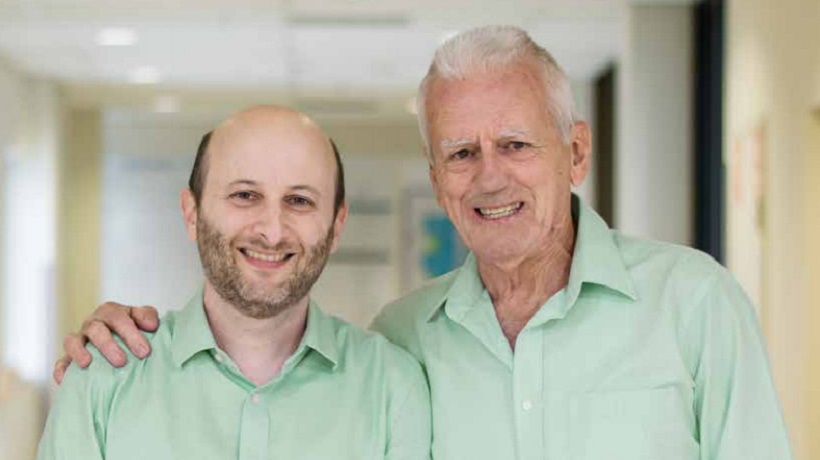 Associate Professor David Ziegler and Col Reynolds are smiling together and wearing matching green shirts; the interior of a building is behind them.