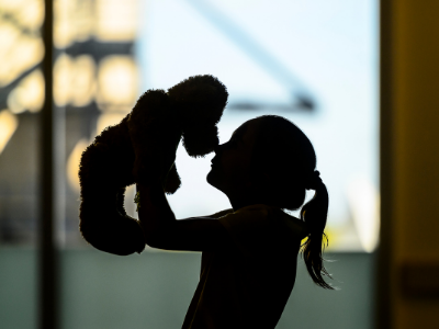 A backlit picture with the silhouette of a girl holding a teddy bear up and close to her face.