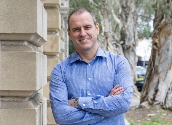 Doug Cunningham smiles with arms folded confidently, behind him is a building and some trees.