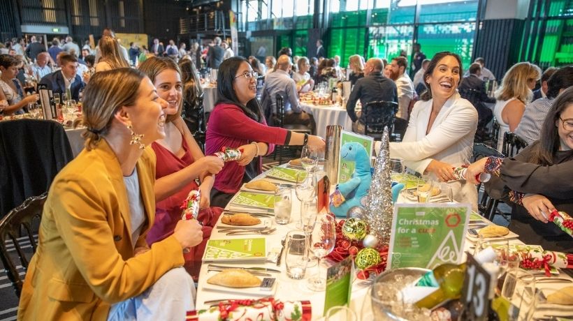 A function room is bustling with smiling people sitting at tables dressed with festive table-settings.