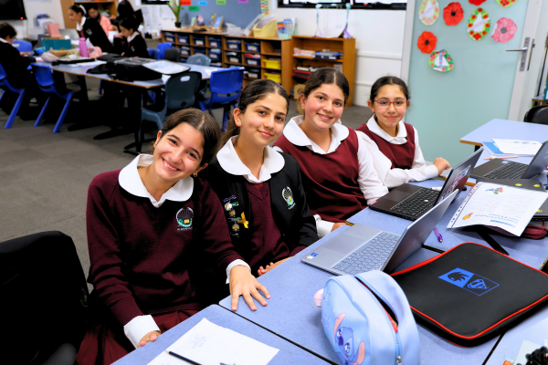 Four school girls sitting at a table in a classroom, smling at the camera, with laptops open on their desks infront of them.