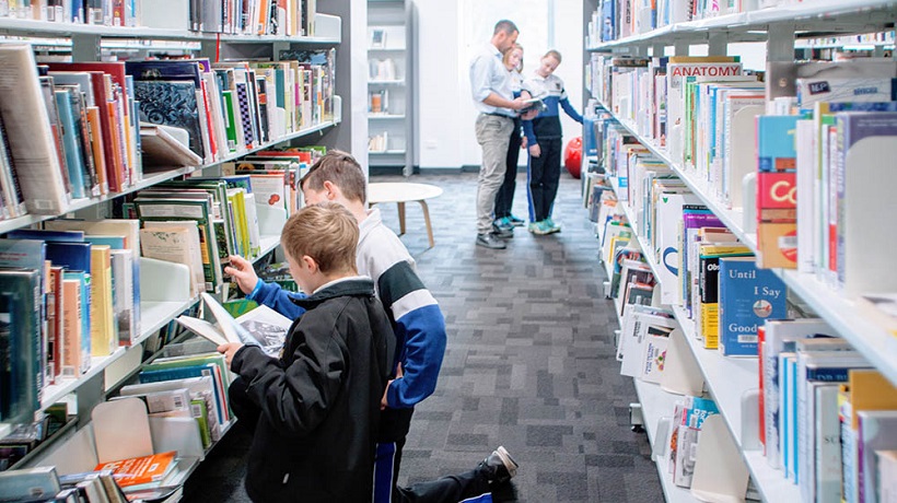 Interior of a library; two children crouch in the foreground, looking at books they have found on the shelves. In the background, a teacher shows another book to two more students.