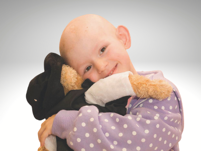 A small child, whose hair has fallen out during treatment, smiles while cuddling a TKCP teddy bear.
