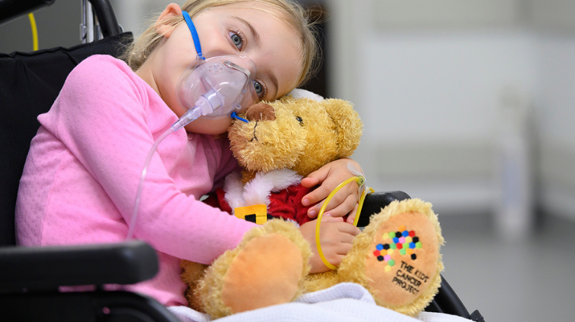 A little girl sitting in a wheelchair and wearing an oxygen mask cuddles her TKCP teddy bear for comfort.