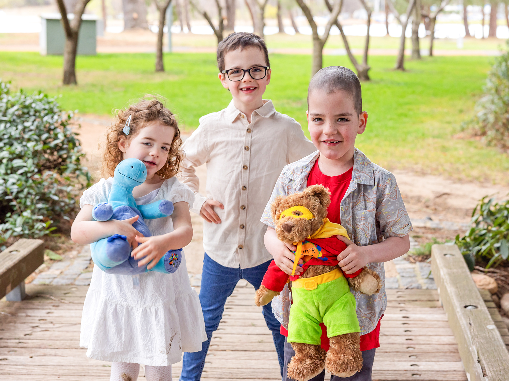A little girl sitting in a wheelchair and wearing an oxygen mask cuddles her TKCP teddy bear for comfort.
