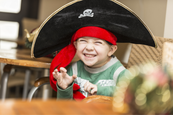A little boy excitedly poses while wearing a very large pirate hat with a big red sash around his forehead.