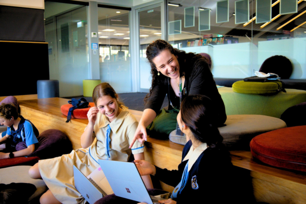 A teacher a two students laughing and smiling, while the teacher points at one of the students' laptop screens in a classroom.